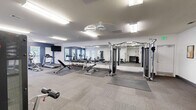 Located at 196 Jeff Rd NW in Huntsville, Alabama, this fitness center offers a comprehensive workout facility spread across a single floor with two dedicated exercise rooms. The space is designed with functionality and comfort in mind, featuring neutral-toned carpet flooring complemented by protective rubber mat sections beneath the equipment.

The main exercise room is generously sized, bathed in bright fluorescent overhead lighting that ensures a well-lit environment throughout. A large wall-mounted mirror spans a significant portion of one wall, enhancing the sense of openness and allowing users to monitor their form during workouts. A wall-mounted flat-screen television adds convenience for cardio sessions.

Equipment is thoughtfully arranged throughout the space and includes multiple treadmills, an elliptical trainer, a stationary bike, a cable machine, a leg press machine, a weight bench, and a dumbbell rack stocked with a full range of free weights and medicine balls. A secondary exercise room provides additional strength training stations and cable equipment.

Natural light filters in through several windows fitted with white blinds, some of which offer views of an outdoor pool area. Accent walls in a deep navy blue provide a modern, energizing aesthetic against the predominantly white walls. The facility also includes ceiling fans and safety amenities such as a fire extinguisher, making it a practical and inviting space for a complete fitness routine.