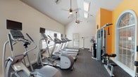 The Crowne Pointe Fitness Center is a thoughtfully designed workout facility housed within a single-floor space distinguished by soaring vaulted ceilings that create an open, airy atmosphere. Skylights and large windows fitted with blinds allow natural light to fill the room, while ceiling fans maintain airflow throughout workouts. The flooring throughout is durable rubber gym matting, practical for both cardio and strength training activities.

The cardio section is well-appointed, featuring a lineup of Precor treadmills and elliptical trainers, each equipped with individual monitor screens for entertainment during exercise. A stair climber and stationary bike round out the cardiovascular offerings. On the strength training side, the facility provides a multi-station cable and resistance machine, along with a comprehensive free weight area stocked with a full rack of dumbbells and fitness accessories such as exercise balls.

Full-length mirrors line one wall, visually expanding the space and allowing users to monitor their form. A bold orange accent wall adds a vibrant, energizing touch to the otherwise neutral palette of white and gray. A glass-paned interior door connects to an additional exercise area, which includes a treadmill and a scale. The overall environment is clean, organized, and well-maintained, offering a complete fitness experience.