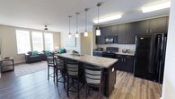 This well-appointed single-floor unit offers a thoughtfully designed open-concept layout that seamlessly connects the kitchen, dining, and living areas. The kitchen is a standout feature, showcasing dark espresso cabinetry extending to the ceiling, granite countertops, and a full suite of black appliances including a refrigerator, electric range, and built-in microwave. A large central island with a built-in sink and bar seating serves as both a functional workspace and a social hub, illuminated by stylish pendant lighting overhead. The living room is bright and welcoming, with large windows that invite natural light and a ceiling fan overhead. Dark hardwood-style flooring runs throughout the common areas, complementing the unit's contemporary aesthetic. Multiple bedrooms offer carpeted floors, individual windows with blinds, ceiling fans, and dedicated desk and chair setups — ideal for focused study or work. The bathrooms are generously sized, each featuring dual-sink vanities topped with granite, dark cabinetry with brushed nickel hardware, wood-style flooring, and a shower-tub combination. The consistent use of dark cabinetry, granite surfaces, and neutral wall tones creates a cohesive, modern atmosphere throughout the entire space.