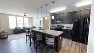 This well-appointed single-floor unit offers a thoughtfully designed open-concept layout that seamlessly connects the kitchen, dining, and living areas. The kitchen is a standout feature, showcasing dark espresso cabinetry extending to the ceiling, granite countertops, and a full suite of black appliances including a refrigerator, electric range, and built-in microwave. A large central island with a built-in sink and bar seating serves as both a functional workspace and a social hub, illuminated by stylish pendant lighting overhead. The living room is bright and welcoming, with large windows that invite natural light and a ceiling fan overhead. Dark hardwood-style flooring runs throughout the common areas, complementing the unit's contemporary aesthetic. Multiple bedrooms offer carpeted floors, individual windows with blinds, ceiling fans, and dedicated desk and chair setups — ideal for focused study or work. The bathrooms are generously sized, each featuring dual-sink vanities topped with granite, dark cabinetry with brushed nickel hardware, wood-style flooring, and a shower-tub combination. The consistent use of dark cabinetry, granite surfaces, and neutral wall tones creates a cohesive, modern atmosphere throughout the entire space.