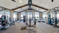The Elevate Fitness Center is a thoughtfully designed, single-floor facility that combines architectural character with a full suite of exercise equipment. The space is defined by its striking vaulted ceiling with exposed dark wood beams, which lend a warm, rustic-modern aesthetic to the interior. Large glass doors and expansive windows line the perimeter walls, flooding the gym with natural light and providing pleasant views of the surrounding greenery outdoors. Natural stone accent columns add a distinctive architectural texture throughout the space.

The main workout area is generously proportioned, providing ample room for movement between equipment stations. A row of modern treadmills and an elliptical trainer are positioned along the windowed wall, allowing users to exercise with an outdoor view. The strength and functional training zone includes a multi-function cable and functional trainer system, a heavy punching bag, battle ropes, a weight bench, and a selection of additional strength machines. Full-length mirrors are strategically placed to enhance the sense of space and support proper form during workouts.

Ceiling fans are installed throughout to ensure comfortable airflow. Entry is through double glass doors framed in matte black, with a clearly posted facility rules sign. The overall ambiance is bright, airy, and motivating — a polished fitness environment suited for a wide variety of training goals.