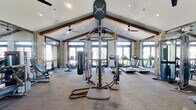 The Elevate Fitness Center is a thoughtfully designed, single-floor facility that combines architectural character with a full suite of exercise equipment. The space is defined by its striking vaulted ceiling with exposed dark wood beams, which lend a warm, rustic-modern aesthetic to the interior. Large glass doors and expansive windows line the perimeter walls, flooding the gym with natural light and providing pleasant views of the surrounding greenery outdoors. Natural stone accent columns add a distinctive architectural texture throughout the space.

The main workout area is generously proportioned, providing ample room for movement between equipment stations. A row of modern treadmills and an elliptical trainer are positioned along the windowed wall, allowing users to exercise with an outdoor view. The strength and functional training zone includes a multi-function cable and functional trainer system, a heavy punching bag, battle ropes, a weight bench, and a selection of additional strength machines. Full-length mirrors are strategically placed to enhance the sense of space and support proper form during workouts.

Ceiling fans are installed throughout to ensure comfortable airflow. Entry is through double glass doors framed in matte black, with a clearly posted facility rules sign. The overall ambiance is bright, airy, and motivating — a polished fitness environment suited for a wide variety of training goals.