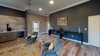 This single-floor communal lounge is thoughtfully designed to serve as both a relaxing retreat and a functional workspace. The open layout is anchored by wood-look flooring, crown molding, and ceiling fans that contribute to a polished yet comfortable atmosphere. Natural light filters in through multiple windows fitted with white blinds, brightening the warm, neutral-toned walls throughout the space.

The lounge is divided into two distinct zones. The primary seating area features a rich cognac leather sofa paired with plush gray accent chairs, all arranged atop a vibrant gold and blue patterned area rug. Bold framed artwork, including a dynamic equestrian painting, adds visual character to the walls. Adjacent to this area, a sleek kitchenette is set against a stacked stone accent wall and includes cabinetry, a sink, a microwave, a beverage refrigerator, and a coffee station—ideal for resident use.

On the opposite end of the room, a deep navy accent wall sets a dramatic backdrop for a long, built-in bar-height workstation with seating for multiple users. A decorative world map artwork anchors this zone, reinforcing a sophisticated, design-forward aesthetic. Additional seating nearby creates a flexible, multi-purpose environment suited for socializing or focused work.