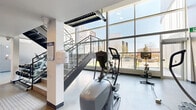 Located at 1792 Pembina Highway in Winnipeg, Manitoba, this well-appointed fitness centre offers a generously sized, open-concept layout spread across a single floor. The space is defined by its striking floor-to-ceiling glass curtain wall, which floods the interior with abundant natural light and provides sweeping views of the surrounding Pembina Highway corridor. The high ceilings and expansive glazing create an airy, energizing atmosphere ideal for exercise. A sleek glass-and-steel staircase serves as both a functional and architectural focal point, adding a modern, industrial edge to the interior. The flooring throughout is a durable, smooth grey surface suited to high-traffic fitness use. The cardio zone is comprehensively equipped, featuring multiple elliptical trainers, treadmills, stationary and spin bikes, and rowing machines. A dedicated strength and free weights area provides dumbbell racks and additional resistance training equipment. Recessed lighting supplements the natural daylight, ensuring consistent illumination throughout. The clean white walls, structured columns, and minimalist aesthetic contribute to a professional and motivating environment. A water station and informational displays are also present, rounding out the practical amenities of this modern, well-designed fitness facility.