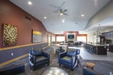 This single-floor space is thoughtfully designed to accommodate both relaxed gatherings and functional events. The entry area sets a welcoming tone with neutral gray walls, patterned carpet tiles, and black leather seating arranged alongside eclectic artwork, including a striking cow painting. Natural light filters in through wood-trimmed windows, and a glass-panel door connects the space to the outdoors.

The great room is the architectural centerpiece, featuring soaring vaulted ceilings, a bold burgundy accent wall, and a wall-mounted television above a sleek tile fireplace. Clerestory windows line the upper walls, flooding the room with abundant natural light. Comfortable seating is arranged in a conversational grouping, complemented by a ceiling fan and recessed lighting. The space transitions seamlessly into a dining area with a large wood table.

The kitchen is well-appointed with rich dark cabinetry, granite countertops, a mosaic tile backsplash, and stainless steel appliances including a full-size refrigerator. An expansive island with seating for multiple guests serves as a practical and social hub. Light wood-look flooring ties the kitchen and dining areas together, creating a cohesive and inviting atmosphere throughout.