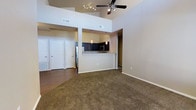 This single-story space is defined by its dramatic vaulted ceilings, which lend an expansive, airy quality to the open-concept layout. The living area is finished with plush carpet underfoot, while the adjacent entryway and hallway transition to hardwood-style flooring, creating a natural visual distinction between zones. A ceiling fan with integrated lighting anchors the upper space, complementing a track lighting fixture that adds a modern touch.

The kitchen sits at the heart of the space, framed by a peninsula-style breakfast bar that separates it from the living area while maintaining an open sightline. Dark cabinetry contrasts with light countertops, and stainless steel appliances, including a refrigerator and built-in microwave, add a sleek, contemporary finish. Pendant lights hang above the peninsula, providing both task lighting and visual interest.

Beyond the kitchen, a hallway leads to additional rooms, with multiple white-paneled doors visible in the background. HVAC vents are positioned throughout, ensuring consistent climate control. The neutral color palette of warm beige and white walls creates a versatile, welcoming ambiance throughout the space.