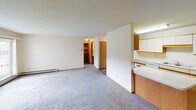 This single-floor residential unit offers a practical and well-organized layout designed for comfortable everyday living. The galley-style kitchen is efficiently arranged with white cabinetry accented by warm wood trim, ample counter space, a double stainless steel sink, a dishwasher, an electric range, and stone-patterned tile flooring beneath a bright overhead light fixture. The kitchen connects seamlessly to a generously sized living area, which features soft carpeting, a wall-mounted air conditioning unit, a baseboard heating system, and a sliding glass door that opens to an outdoor balcony. An open walkway leads to additional storage and closet space, adding to the unit's overall functionality. The bedrooms are carpeted and well-lit, each with wood-trimmed windows that bring in natural light and baseboard heaters for year-round comfort. The bathroom is thoughtfully appointed with a vanity featuring wood cabinetry and a light countertop, a large wall mirror, a tub-shower combination, stone-patterned tile flooring, and grab bars for added accessibility. Textured ceilings are consistent throughout the unit, contributing to a cohesive interior aesthetic. Overall, the space presents a clean, neutral palette with warm wood accents that unify each area.
