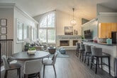 Located at 103 Little Bay Ave in Yorktown, Virginia, this single-story home offers an open, airy layout with thoughtfully designed living spaces filled with abundant natural light. The living room serves as the heart of the home, boasting dramatic vaulted ceilings that soar to impressive heights, framed by a striking arched window that floods the space with daylight. A full-height painted brick fireplace anchors the room, providing a classic focal point that complements the warm, neutral palette throughout. Dark hardwood flooring adds richness and continuity across the open-plan living and dining areas. The kitchen is well-appointed with light wood cabinetry, dark countertops, stainless steel appliances, and large windows that invite in natural light while offering views of the surrounding landscape. A dedicated home office area is enclosed with an elegant art-glass entry door and features a continuous granite-topped built-in desk along one wall, flanked by multiple windows with wood blinds — creating a bright, functional workspace. A tray ceiling detail lends architectural interest to this room. Throughout the home, crisp white trim, soft neutral wall tones, and generous window placement create a cohesive, welcoming ambiance suited to both relaxed living and productive daily routines.