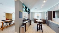 Located at 1227 Ashes Dr in Wilmington, NC, this thoughtfully designed single-floor amenity space combines modern aesthetics with functional gathering areas. Light hardwood flooring flows throughout, unifying the various zones and lending a warm, cohesive feel to the interior.

The open social hub at the heart of the space features a wet bar with dark countertops, a stainless steel under-counter refrigerator, and bold teal open shelving, providing both practicality and a striking visual accent. Adjacent bar-style seating with leather and wood stools lines the counter, while a long marble-topped communal dining table beneath a sculptural multi-globe pendant light anchors the dining area. French doors offer direct views and access to an outdoor pool courtyard, flooding the space with natural light.

The lounge area showcases a geometric patterned accent wall, woven lounge chairs with mint and coral cushions, and curated artwork, creating an inviting, resort-like atmosphere. A mounted television and built-in cabinetry add functionality. A cozy seating nook with a tufted leather bench and wall sconces provides a quieter retreat.

A dedicated conference room offers a dark stone table, cognac leather chairs, and a wall-mounted display, suitable for private meetings or focused work.