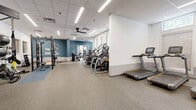 This expansive fitness center offers a comprehensive, single-floor layout designed to accommodate a wide range of workout needs. The space is thoughtfully organized into distinct functional zones, beginning with a dedicated cardio area lined with treadmills and elliptical machines positioned near windows that allow natural light to filter in. Ceiling fans and a drop tile ceiling with recessed linear lighting ensure the space remains comfortable and well-illuminated throughout.

The strength training zone features a Matrix power rack fully loaded with Olympic weight plates, a TRX suspension training station, a full dumbbell rack, and multiple adjustable benches arranged on durable rubber flooring. A blue-toned accent area provides a designated space for free weight and functional training. Patterned tile wainscoting along the lower walls adds a distinctive architectural detail that runs throughout the main workout area.

A separate studio or flex room with light hardwood flooring, starburst pendant light fixtures, and bold teal accent walls creates an inviting space for stretching, yoga, or small group sessions. Stability balls, a mat station, and an interactive fitness mirror round out the studio's offerings. Motivational wall graphics and a cohesive modern color palette give the entire fitness center an energetic yet polished ambiance suited for focused training.