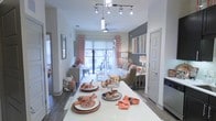 This thoughtfully designed single-floor residence showcases a modern open-concept layout that effortlessly connects the kitchen, dining, and living spaces. The kitchen features a sleek white island that doubles as a dining surface, illuminated by a contemporary track lighting fixture overhead, lending both functionality and visual interest to the space. Rich hardwood-style flooring runs throughout, grounding the interior with warmth and continuity. The dining area is elegantly staged with layered place settings and warm coral and copper accents that carry cohesively into the living room beyond. The living space is anchored by a cozy seating arrangement, complemented by an accent wall in a deep charcoal tone that adds depth and sophistication. Large windows dressed in coral-patterned curtains and light-filtering roller shades flood the space with natural light while offering a view to an outdoor balcony or patio area. Decorative artwork, ambient table lamps, and carefully curated accessories contribute to the warm, inviting atmosphere. The overall ambiance strikes a balance between stylish and livable, making it an appealing and well-appointed residential space.