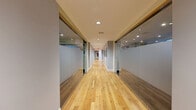 Located at 205 Church Street in the heart of New Haven, CT, this well-appointed fitness facility spans a generous single floor and is organized into four distinct exercise rooms, each designed to serve a specific training purpose. A sleek, light-filled corridor with warm hardwood flooring and full-length glass partition walls connects the spaces, creating an open and transparent flow throughout the facility. The first exercise room functions as a dedicated studio space, featuring polished hardwood floors, floor-to-ceiling mirrors, and an assortment of free weights including dumbbells, kettlebells, and stability balls — ideal for group classes or personal training sessions. A second room is outfitted with cardio equipment, including treadmills, elliptical machines, and a stationary bike, all positioned beneath recessed lighting with natural light filtering in through street-facing windows. A third room is dedicated to strength training, housing a variety of plate-loaded machines and cable systems on durable rubber flooring, with windows providing natural light and ventilation. The fourth room completes the circuit with additional free weights, a multi-station cable machine, and mirrored walls. Recessed lighting throughout ensures consistent, well-lit conditions across all areas, and the overall design reflects a clean, modern aesthetic suited for a professional fitness environment.