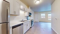 This single-floor unit offers a clean, modern design characterized by light wood-look flooring, neutral warm-toned walls, and consistent flush-mount lighting throughout. The kitchen is outfitted with white shaker-style cabinetry featuring brushed nickel hardware, white countertops, a stainless steel double-basin sink, and a full suite of stainless steel appliances including a range, over-the-range microwave, dishwasher, and refrigerator. The layout allows the kitchen to flow seamlessly into an open living area, which benefits from natural light provided by a large window overlooking an outdoor landscape. A hallway connects the main living space to the private rooms, with a dedicated entry door visible from the kitchen area. The bedroom is a well-proportioned, open room with its own window offering outdoor views and ample wall space for flexible furniture arrangements. The bathroom is designed with accessibility in mind, featuring stainless steel grab bars, a white shaker-style vanity with an integrated sink, a large wall mirror, and light wood-look flooring that ties cohesively to the rest of the unit. Overall, the space presents a bright, functional, and accessible living environment.