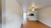 This two-story space is designed with a practical and clean aesthetic, featuring a neutral color palette of whites and soft beiges throughout. The main level showcases warm wood-style flooring that flows through the open living area and into the kitchen. The living space is anchored by a ceiling fan light fixture and includes built-in open shelving along one wall, offering functional display and storage. A sliding glass door with vertical blinds fills the area with natural light and provides outdoor access.

The galley-style kitchen is well-appointed with white cabinetry extending to the ceiling, granite-style laminate countertops, a double basin sink, and black appliances including an electric range and refrigerator. A peninsula counter separates the kitchen from the living area, creating a natural divide while maintaining an open feel.

A staircase leads to the upper level, where soft carpet continues throughout. The upper floor features an open landing with a decorative railing and access to additional rooms. A bathroom on this level includes a vanity with a granite-style countertop, a medicine cabinet mirror, and a separate tub and shower area with wood-style flooring. Ceiling fans are present in the upper-level rooms, which benefit from natural light through windows.