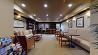 This thoughtfully designed single-floor communal space functions as both a library and multipurpose lounge, blending several distinct zones into one cohesive environment. The layout opens with an entry foyer featuring frosted-glass double doors flanked by wall sconces, a tall potted palm, and a wooden bench with a literature rack — establishing an inviting, polished first impression.

The main lounge area centers on a cream upholstered sofa and matching armchair arranged around a wooden coffee table stacked with books, set against built-in bookshelves filled with reading material. Framed artwork and wall-mounted sconces add warmth to the neutral-toned walls, while recessed lighting and patterned carpeting throughout reinforce the refined atmosphere.

A dedicated study area features two computer workstations set within dark wood built-ins, surrounded by shelving stocked with books and decorative objects. Windows fitted with horizontal blinds provide controlled natural light to this workspace.

Along one wall, a banquette-style seating section with upholstered bench seating, wooden café tables, and cushioned arm chairs offers a casual dining or social gathering spot, accented by nautical-themed framed prints and sconce lighting.