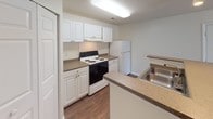 This single-floor residential unit offers a practical and well-organized layout designed for comfortable everyday living. The kitchen is a standout feature, outfitted with crisp white cabinetry — both upper and lower — providing ample storage space. Laminate countertops in a neutral tone complement the white aesthetic, while an electric range with coil burners and a full-size refrigerator supply essential cooking and food storage needs. A double-basin stainless steel sink is conveniently positioned at a peninsula-style counter, adding workspace and a subtle open connection to the adjoining living area. Rich wood-look flooring runs throughout the kitchen, adding warmth and easy maintenance to the space. The living area is generously sized, finished with plush neutral carpeting that creates a soft, inviting atmosphere. Natural light filters in through a large window fitted with horizontal blinds, while a sliding glass patio door — covered with vertical blinds — extends the space visually and provides direct outdoor access. The bathroom is cleanly appointed with a full-size soaking tub and shower combination, chrome fixtures, and matching wood-look flooring that ties back to the kitchen aesthetic. White walls and a textured ceiling are consistent throughout, lending the unit a cohesive, bright, and move-in-ready feel.