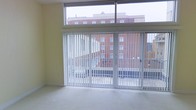 This single-floor space is defined by its bright, open character and modern architectural details. The most prominent feature is the expansive wall of floor-to-ceiling windows combined with a wide sliding glass door, which floods the interior with abundant natural light and provides sweeping views of the surrounding urban environment, including a notable brick building across the way. Vertical blinds offer flexible light and privacy control throughout the day. The sliding door opens directly to a balcony enclosed with metal railings, seamlessly connecting the interior to the outdoors. The interior walls are finished in a soft, warm yellow tone that complements the light-toned flooring, contributing to an airy and welcoming ambiance. A transom-style window runs along the upper portion of the window wall, further enhancing the sense of openness and height. The overall design reflects a clean, contemporary aesthetic with an emphasis on natural light, urban connectivity, and functional simplicity. The space presents a versatile, uncluttered layout suited for a variety of uses.