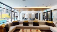 This contemporary single-floor space is thoughtfully designed to accommodate a variety of social and recreational activities, blending modern architecture with curated interior design throughout. The entry corridor features warm wood-paneled ceilings with cove lighting, large glass doors, and a welcoming atmosphere that flows into multiple distinct zones.

The main lounge area centers around a generously sized sectional sofa in neutral tones, accented with mustard and charcoal throw pillows, arranged over an abstract area rug. Adjacent dining areas offer round tables paired with sleek dark chairs, illuminated by cascading pendant light fixtures. A striking three-dimensional floral wall sculpture serves as a notable artistic focal point in the dining zone.

A well-appointed wet bar features a white quartz countertop, stainless steel sink, integrated shelving with decorative greenery, and a mounted flat-screen television. Nearby, a bold blue upholstered banquette nook provides an intimate seating option beside warm hardwood flooring.

The game room stands out with a rustic wood-framed billiards table, an industrial overhead light fixture, mounted televisions, and large-scale abstract murals that energize the space. Floor-to-ceiling windows and glass partitions throughout ensure natural light permeates every area.