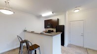 This single-floor space showcases a well-appointed kitchen that serves as a central feature of the layout. The kitchen is designed with rich, dark espresso-toned cabinetry that provides ample upper storage, complemented by sleek black appliances including a refrigerator, microwave, and stove. A peninsula-style breakfast bar extends from the kitchen, offering a laminate countertop surface and accommodating bar-height seating with metal and wood stools — making it a practical space for casual dining or entertaining. Crown molding along the ceiling adds a refined architectural detail, lending the space a polished, finished look. The flooring features neutral-toned tile, contributing to a clean and cohesive aesthetic throughout. Recessed and flush-mount lighting fixtures provide consistent illumination across the kitchen and adjacent areas. A hallway visible beyond the kitchen leads to additional rooms, suggesting a functional and straightforward floor plan. The white walls and light-colored ceiling enhance brightness, creating an open and airy atmosphere throughout the space.