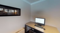 This single-floor computer center is a purpose-built space designed to provide residents or guests with essential technology access in a comfortable, well-maintained setting. The room features clean, neutral-toned walls accented by elegant crown molding along the ceiling, lending a polished and refined character to what is fundamentally a functional workspace.

The workstation area is equipped with a desktop computer, monitor, keyboard, and mouse, all arranged on a streamlined dark-framed desk with a light-colored work surface. A wall-mounted flat-screen display adds an additional informational or media resource to the space. A framed piece of wall art contributes a touch of decor, softening the utilitarian nature of the room.

A printer and additional desk surface are also present, supporting document printing needs. A prominently displayed "Computer Center Rules" sign — attributed to Cedar Edlen Management — outlines policies including restrictions on food and beverages, age requirements, download limitations, print guidelines, and session time limits, ensuring orderly and fair use of the facility.

The space is accessible through a dark-framed doorway and benefits from natural light through a window, maintaining a bright and inviting atmosphere. The overall design balances practicality with a clean aesthetic, making it a well-organized resource for users seeking reliable computer and printing access.