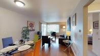 This single-floor space combines a dining area and living room in an open-concept layout, offering a functional and welcoming atmosphere. The hardwood flooring runs throughout, providing warmth and visual continuity between the two zones. In the dining area, a round table is paired with vibrant green and orange chairs, adding a lively pop of color against the neutral walls. A flat-screen television on a low media stand anchors one side of the space, while a large window along the far wall allows generous natural light to fill the room. The living area is furnished with a dark leather sofa and armchair, arranged around a glass-top coffee table, creating a comfortable and cohesive seating arrangement. A stylish floor lamp and table lamp provide warm ambient lighting. Framed artwork and a large canvas painting decorate the walls, contributing character and visual interest. A potted plant near the dining area introduces a natural element. The overall ambiance is bright, modern, and inviting, with a clean, well-organized layout that maximizes the sense of space throughout.