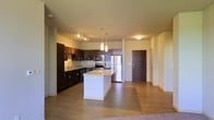 This single-floor space showcases a thoughtfully designed open-concept layout that seamlessly connects the kitchen and living areas, creating a sense of spaciousness and flow. The kitchen is a standout feature, anchored by a central island with a light granite countertop and elegant pendant lighting overhead. Rich, dark espresso cabinetry lines the walls, complemented by matching granite countertops along the perimeter and a full suite of stainless steel appliances, including a refrigerator, range, and built-in microwave. A track lighting system above the main cabinetry run provides additional task lighting, while a flush ceiling fixture brightens the entry area. The flooring transitions from smooth hardwood-style planks in the kitchen and entryway to carpeting in the living space, adding warmth and texture. The entry door is finished in a dark wood tone, contributing to the cohesive, contemporary aesthetic carried throughout the unit. Clean, light-toned walls and generous ceiling height lend the space an airy, modern quality. The overall design balances functionality with style, making the kitchen the true centerpiece of this well-appointed, single-level home.