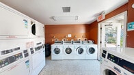 This shared laundry facility is a compact, single-room space designed for efficient, high-volume use. The room is immediately distinguished by its bold terracotta-orange accent wall, which adds a warm, inviting contrast to the predominantly white commercial equipment that lines the perimeter. A row of front-loading commercial washers — branded Speed Queen — is arranged along the main wall, each equipped with digital controls and clearly labeled for ease of use. Along the side wall, a bank of stacked commercial dryers provides ample drying capacity, with individual units numbered for user convenience. The terrazzo flooring offers a durable, easy-to-clean surface well-suited to the demands of a shared laundry environment. Overhead, a flush-mounted fluorescent light fixture ensures bright, even illumination throughout the space. Wall-mounted sconce lights above the washers provide additional task lighting. An operational instructions board and payment information signage are posted near the entry door, which features a glass panel allowing natural light to filter in from outside. A ceiling-mounted HVAC vent supports proper ventilation, keeping the space comfortable during use. The layout is straightforward and functional, maximizing the available floor area to accommodate multiple users simultaneously.