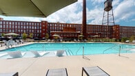 This outdoor amenity space in South Carolina offers a thoughtfully designed environment that blends industrial heritage with modern recreational comfort. The centerpiece is a generously sized resort-style swimming pool with crystal-clear water, surrounded by a wide concrete pool deck lined with sleek chaise lounges and umbrella-shaded seating. A striking historic red brick mill building with a towering smokestack forms an impressive backdrop, adding character and architectural depth to the setting.

Adjacent to the pool, a covered pavilion constructed with exposed wood timber framing and brick accent walls provides a shaded retreat ideal for gathering and relaxation. The pavilion is equipped with comfortable outdoor lounge furniture, Adirondack chairs, a fireplace, and wall-mounted televisions, creating an inviting atmosphere for both casual and social occasions. String lights overhead enhance the ambiance during evening hours.

Beyond the main pool area, additional amenities include outdoor grilling stations and a spa, all set within a well-maintained patio environment. The open-air design allows for abundant natural light and fresh air throughout, while mature trees along the perimeter provide natural greenery. The combination of historic architectural surroundings and contemporary outdoor furnishings makes this space a distinctive and functional amenity destination.