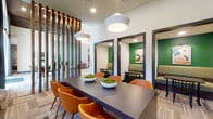 This single-floor amenity space is thoughtfully designed to accommodate a variety of social and recreational activities, offering distinct zones that flow seamlessly into one another. A dramatic vertical wood slat partition anchors the layout, serving as a visual divider between areas while adding a warm, natural texture to the contemporary interior.

The dining area features a long dark communal table surrounded by vibrant orange upholstered chairs, lit by oversized pendant lights. Adjacent to it, a series of intimate booth-style nooks are set into alcoves with bold green accent walls, striped bench seating, and curated artwork — ideal for small gatherings or focused conversations.

The kitchen area is highlighted by a striking patterned tile backsplash, dark cabinetry, and a generous white marble-topped island with bar stools, complemented by sculptural wire pendant fixtures above.

The lounge zone features soaring ceilings, plush seating arranged around a rustic wood coffee table, and a large wall-mounted television — perfect for casual viewing or socializing. Nearby, a full-size billiards table sits beneath a vaulted wood-paneled ceiling with circular statement lighting, completing the entertainment offerings of this well-appointed communal space.