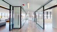 This contemporary single-floor commercial space showcases a refined, modern design built around transparency, light, and open-plan functionality. The layout is anchored by a welcoming entrance lobby featuring large floor-to-ceiling windows, polished large-format tile flooring, and elegant glass railings, offering sweeping outdoor views and an immediate sense of openness.

Throughout the space, full-height glass partition walls with dark aluminum framing create distinct zones while maintaining visual connectivity and allowing natural light to permeate every corner. A spacious lounge area serves as a central gathering point, furnished with generous modular sofas, a rounded coffee table, and wall-mounted display screens — ideal for informal meetings or relaxation. High-top tables and bar seating along the window line provide additional collaborative spots with panoramic views of the surrounding landscape.

The open-plan areas are bright and versatile, benefiting from expansive window walls fitted with roller shades for adjustable light control. Recessed linear lighting supplements the natural light throughout. Cylindrical white structural columns add architectural character without obstructing the flowing layout. The overall ambiance is clean, professional, and inviting — a well-appointed commercial environment designed to support both focused work and collaborative engagement.