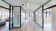 This contemporary single-floor commercial space showcases a refined, modern design built around transparency, light, and open-plan functionality. The layout is anchored by a welcoming entrance lobby featuring large floor-to-ceiling windows, polished large-format tile flooring, and elegant glass railings, offering sweeping outdoor views and an immediate sense of openness.

Throughout the space, full-height glass partition walls with dark aluminum framing create distinct zones while maintaining visual connectivity and allowing natural light to permeate every corner. A spacious lounge area serves as a central gathering point, furnished with generous modular sofas, a rounded coffee table, and wall-mounted display screens — ideal for informal meetings or relaxation. High-top tables and bar seating along the window line provide additional collaborative spots with panoramic views of the surrounding landscape.

The open-plan areas are bright and versatile, benefiting from expansive window walls fitted with roller shades for adjustable light control. Recessed linear lighting supplements the natural light throughout. Cylindrical white structural columns add architectural character without obstructing the flowing layout. The overall ambiance is clean, professional, and inviting — a well-appointed commercial environment designed to support both focused work and collaborative engagement.