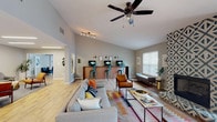 Located at 100 Northwoods Dr in Cary, NC, this single-floor community clubhouse offers a thoughtfully designed gathering space with a vibrant, eclectic aesthetic rooted in mid-century modern style. The main lounge is expansive and airy, featuring soaring vaulted ceilings, ceiling fans, and light wood-look flooring that flows throughout. A dramatic floor-to-ceiling geometric-patterned fireplace serves as a striking focal point, complemented by a comfortable sectional sofa, leather accent chairs, and colorful kilim-style area rugs. Natural light pours in through multiple windows, enhancing the warm, welcoming ambiance. A built-in business center with computer stations and teal industrial bar stools lines one wall, offering a functional workspace within the social setting. Adjacent seating areas provide additional lounge space with geometric side tables and curated artwork on the walls. A dedicated game room includes a pool table and shuffleboard for recreation. The full kitchen is well-appointed with white cabinetry, stainless steel appliances, a mosaic tile backsplash, pendant lighting, and ample counter space. A long communal high-top wooden bar table with industrial metal stools offers casual dining or co-working. The space seamlessly balances comfort, style, and utility, making it an ideal hub for community gathering and relaxation.