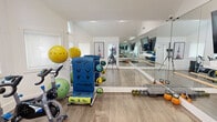 This private fitness studio is thoughtfully designed to deliver a comprehensive workout experience within a compact, efficient footprint. The space is defined by striking vaulted ceilings that lend an airy, open feel, while an arched transom window alongside multiple standard windows floods the room with abundant natural light. The warm wood-look flooring provides both comfort and durability underfoot.

A full wall of floor-to-ceiling mirrors dominates one side of the studio, visually expanding the space and supporting form-checking during exercise. Mounted on the mirror wall, a large flat-screen television enables guided workout sessions or entertainment. A ballet barre runs along the mirrored wall, catering to barre and flexibility training.

The studio is well-stocked with a diverse range of equipment, including Stages cycling bikes for indoor cycling, a rack of colorful dumbbells, kettlebells, medicine balls, stability balls, a SmartMat exercise mat station, and step platforms. An adjacent room, accessible through a glass-paned interior door, houses additional cardio machines including a treadmill and elliptical trainer.

Crisp white walls and recessed lighting complete the clean, modern aesthetic. The overall ambiance is bright, motivating, and professionally appointed, making this studio an ideal setting for personal training, group classes, and multi-discipline fitness routines.