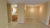 This single-floor space presents a straightforward, functional layout that connects the main living area to the kitchen in an open, semi-divided arrangement. A structural half-wall separates the two zones, providing a degree of definition while maintaining a sense of openness between them.

The living area is finished with light-toned carpeting and neutral beige walls, creating a warm, understated atmosphere. A track light fixture is mounted to the ceiling, providing overhead illumination, while a small pass-through window is built into the dividing wall, adding a subtle architectural detail and allowing light to travel between spaces.

The kitchen features warm-toned wooden cabinetry along the upper and lower walls, paired with a double sink positioned beneath a window that allows natural light into the space. The kitchen floor transitions to hard flooring, offering a practical contrast to the carpeted living area.

White baseboards run throughout, providing clean definition between the walls and flooring. The overall palette of warm beiges and natural wood tones gives the space a cohesive, neutral character well-suited to a variety of furnishing styles.
