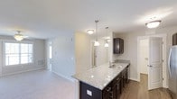 Located at 3400 Briercliff Dr in Greenville, NC, this single-story home offers a thoughtfully designed open-concept layout that connects the kitchen and living areas in a cohesive, functional flow. The kitchen serves as a central highlight, featuring a peninsula with striking granite countertops in a light speckled finish, contrasted elegantly by dark espresso cabinetry with brushed nickel hardware. A stainless steel sink is integrated into the peninsula, which also provides casual seating or prep space. Overhead, pendant lights with polished nickel fixtures hang above the peninsula, adding a modern touch to the space. The kitchen transitions naturally into the adjacent living area, which is finished with neutral-toned carpet, creating a warm and comfortable environment. Light-toned walls throughout contribute to a bright, airy atmosphere. A doorway visible in the background hints at additional rooms beyond, while a window with natural light is visible in the adjoining room, enhancing the overall openness of the home. The combination of hard surface flooring in the kitchen and soft carpet in the living area defines each zone while maintaining visual continuity throughout the single-floor layout.