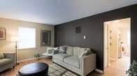 This single-floor space showcases a thoughtfully designed living area that balances modern aesthetics with a welcoming ambiance. The living room serves as the focal point, featuring a striking dark charcoal accent wall that creates a bold contrast against the lighter taupe-toned surrounding walls and crisp white ceiling. This design choice adds depth and a contemporary edge to the room.

Natural light enters through a window fitted with a white blind, brightening the space and complementing the warm-toned hardwood flooring that runs throughout. A light gray sofa anchors the seating area, accessorized with patterned throw pillows in neutral tones. A sleek accent chair and a small side table provide additional seating, while a decorative framed mirror mounted on the wall adds visual dimension.

A textured area rug in neutral tones grounds the seating arrangement, and a dark leather ottoman introduces contrast at the foreground. A small potted plant near the window adds a natural, organic touch. A doorway visible on the right suggests connectivity to additional rooms, contributing to the overall functional flow of this well-appointed space.