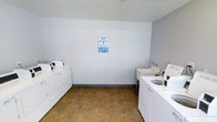 This single-floor laundry room is designed with practicality and functionality in mind, providing a well-organized space for laundry tasks. The room features multiple top-loading washing machines and dryers arranged along one wall, offering ample capacity for handling laundry efficiently. On the opposite side, a set of white utility countertops provides convenient workspace, accompanied by a utility sink ideal for hand-washing or pre-treating garments. The walls are finished in a clean, neutral light grey, contributing to a bright and tidy atmosphere. A posted laundry instruction guide on the main wall ensures users have easy access to operational directions. The flooring consists of durable, earth-toned vinyl tile that is well-suited for a high-traffic utility space and easy to maintain. Overhead, flush-mounted lighting provides consistent, functional illumination throughout the room. The overall layout is straightforward and efficient, maximizing usable floor space while keeping all essential laundry equipment and workspaces easily accessible. The space presents a clean, no-frills environment focused entirely on meeting practical laundry needs.