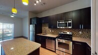 This single-floor residence offers a thoughtfully designed layout that seamlessly connects the kitchen, dining, and living areas. The kitchen is a standout feature, showcasing rich dark cabinetry with sleek hardware, light granite countertops, and a full suite of stainless steel appliances including a refrigerator, range with oven, built-in microwave, and dishwasher. A striking mosaic tile backsplash in warm earth tones adds texture and visual interest, while a central island with a matching granite surface provides additional prep space and casual seating. Vibrant pendant lighting in yellow accents the kitchen, creating a warm and inviting atmosphere. The living area continues the warm aesthetic with wood-style flooring that extends throughout the main spaces, neutral-toned walls, and a ceiling fan that enhances airflow and comfort. A sliding glass door floods the living space with natural light and opens to an outdoor view of greenery. The bathroom features a glass-enclosed walk-in shower with clean white subway tile, chrome fixtures, and a rainfall-style showerhead, offering a crisp, modern finish. The carpeted bedroom area adds a cozy contrast to the hard flooring found elsewhere in the unit, completing this well-balanced and functional living space.