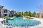 Located in Columbus, Georgia, this outdoor amenity space showcases a beautifully designed resort-style pool and expansive patio area. The centerpiece is a large, freeform swimming pool with a gradual entry, its clear waters reflecting the surrounding natural scenery. The pool's curved edges and stone coping contribute to an elegant, inviting aesthetic.

Surrounding the pool, a generously sized stone-paved patio provides ample room for relaxation and socializing. Numerous sling-style chaise lounges with wood frames are arranged throughout the deck, offering comfortable spots to lounge in the Georgia sunshine. Teal patio umbrellas provide shaded seating areas, adding a pop of color against the neutral stone and dark exterior of the main building.

The backdrop features a striking multi-story structure with a stone and dark-siding exterior, accented by large windows and a covered balcony, lending an upscale architectural presence to the space. Mature evergreen and deciduous trees frame the perimeter, creating a natural privacy buffer and a serene, park-like atmosphere. The combination of refined hardscaping, thoughtful furnishings, and abundant greenery makes this outdoor space a standout feature of the property.