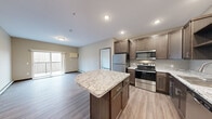 Located in Cold Spring, Minnesota, this single-floor residence offers a thoughtfully designed layout with modern finishes throughout. The open-concept kitchen serves as the heart of the unit, showcasing rich dark-brown cabinetry, a striking granite-topped center island, and a full suite of stainless steel appliances including a refrigerator, electric range, microwave, and dishwasher. Light wood-tone flooring runs through the kitchen and living areas, lending a clean, contemporary feel. The living area is bright and open, with a sliding glass door that leads to a private balcony overlooking a natural, wooded backdrop — ideal for enjoying Minnesota's seasonal scenery. The bedroom is generously sized, finished with plush carpeting and a large window that frames views of the surrounding trees, filling the room with natural light. A spacious walk-in closet features wire shelving and hanging rods for ample organization. The bathroom is well-appointed with dark cabinetry, a white countertop vanity, a large mirror, and wood-look flooring. A dedicated in-unit laundry area includes a full-size washer and dryer with overhead cabinetry and wire shelving for added storage. The layout is efficient and functional, delivering comfort and modern living in a serene setting.
