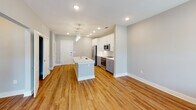This single-floor residence at 5634 McCrimmon Pkwy in Morrisville, NC, offers a clean, modern aesthetic throughout. The open-concept living and kitchen area is anchored by warm hardwood-style flooring and recessed lighting that creates a bright, welcoming atmosphere. The kitchen is well-appointed with white upper cabinetry, gray lower cabinets, stainless steel appliances including a side-by-side refrigerator, electric range, and built-in microwave. A geometric hexagonal tile backsplash adds a stylish detail, while a central island with a sink and pendant lighting serves as a functional focal point for the space.

The bedroom features neutral carpet flooring, soft gray walls, and direct sightlines to the kitchen area. A walk-in closet is outfitted with wire shelving and hanging rods, providing organized, practical storage. An additional carpeted room with two large windows and built-in wire shelving offers flexibility as a secondary bedroom or storage space.

The bathroom is finished with hardwood-style flooring, a lengthy white floating vanity with a light stone countertop, a framed mirror, and a glass-enclosed walk-in shower with floor-to-ceiling tile. The overall design is cohesive, emphasizing clean lines, neutral tones, and functional finishes throughout.