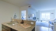 This single-floor residence showcases an open-concept layout that connects the kitchen and living areas in a bright, cohesive design. The kitchen features granite countertops with warm beige and gold tones, providing generous workspace and a stainless steel sink. A raised breakfast bar creates a natural division between the kitchen and the adjoining spaces while maintaining visual openness throughout.

The white walls and light-toned ceilings amplify the abundant natural light that flows in from large windows visible in the living area, creating an airy and welcoming atmosphere. The living room, visible from the kitchen, is carpeted and centers around a fireplace framed by a clean, white mantel, adding a classic focal point to the space. Hardwood-style flooring connects transitional areas, complementing the neutral color palette.

A doorway from the kitchen leads to an additional room, likely a bedroom or office, which features its own natural light source and glimpses of tasteful furnishings. Overall, the space presents a well-lit, neutral, and functional interior with a cohesive design that balances comfort and practicality throughout its single level.