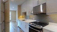 This single-floor space showcases a thoughtfully designed kitchen that serves as a central highlight. The kitchen features a clean, modern aesthetic with flat-front white cabinetry extending to the upper walls, providing generous storage. A white quartz countertop runs the length of the kitchen, offering ample workspace and a bright, polished surface. A gray subway tile backsplash adds subtle texture and contrast against the white cabinetry and counters. Stainless steel appliances include a gas range with multiple burners and a matching range hood overhead, complemented by a built-in dishwasher. A stainless steel undermount sink with a pull-down faucet is conveniently positioned along the countertop. Light wood-tone flooring runs throughout, contributing warmth to the otherwise neutral palette. The layout is efficiently organized, with a corridor visible beyond the kitchen leading further into the space. Recessed lighting illuminates the area, enhancing the overall brightness and open feel of the interior. The combination of functional design and contemporary materials gives this space a clean, move-in-ready character.