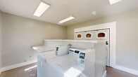 This shared laundry facility is a clean, efficiently designed space that provides all the essentials for a convenient laundry experience. The room is outfitted with a row of top-loading washing machines positioned along one side, complemented by a bank of stacked dryers along the opposite wall, offering ample capacity for multiple users. A continuous stone-topped folding counter runs alongside the washers, providing a practical surface for sorting and folding laundry. The interior is finished with neutral, light-toned walls and smooth, wood-look flooring that is both durable and easy to maintain. Overhead, flush-mounted fluorescent lighting panels ensure the space is brightly and evenly lit. Large, multi-pane windows with dark frames are a standout architectural feature, flooding the room with generous natural light and offering views of the surrounding greenery. A glass-paneled entry door further enhances the brightness of the space. Safety is addressed with a prominently mounted fire extinguisher near the entrance. The overall ambiance is tidy, functional, and welcoming, reflecting a well-maintained communal facility designed to serve residents with ease and practicality.