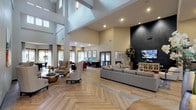 This thoughtfully designed clubroom offers an expansive, open-concept layout highlighted by dramatic soaring ceilings and striking herringbone-patterned hardwood flooring that flows throughout the entire space. The main lounge area is anchored by a large sectional sofa accented with colorful throw pillows, a flat-screen TV mounted against a bold dark feature wall, and a stylish coffee table — creating an inviting entertainment hub. A circular upholstered ottoman serves as a central focal point in the open gathering area, beneath a statement sputnik chandelier that adds a modern, sophisticated touch.

Adjacent to the lounge, a well-appointed business center features multiple desktop workstations along a granite-topped counter, ergonomic seating, and a wall of framed artwork, all bathed in natural light from large windows overlooking the outdoor pool area. Nearby, a cozy dining nook with a round wood table and teal studded chairs is framed by floor-to-ceiling windows and sliding glass doors, offering direct views of the pool and surrounding greenery.

A wet bar and counter-height seating area provide an additional social space at the rear of the room. Throughout, neutral warm tones, layered lighting, and curated décor contribute to a polished yet welcoming ambiance suited for both relaxation and productivity.