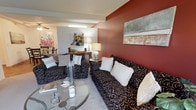 This single-floor residence offers a thoughtfully designed open-concept layout that seamlessly connects the living and dining areas, creating a cohesive and inviting atmosphere. The living area is anchored by a rich, deep red accent wall that adds warmth and visual depth to the space, complemented by neutral tones on the surrounding walls and a textured ceiling that adds dimension overhead. Exposed beams add an architectural touch, lending structure and character to the open floor plan. The living space is furnished with plush, patterned sofas and accent chairs arranged around a round glass coffee table, creating an elegant and comfortable gathering area. Decorative artwork adorns the walls, contributing to a curated and expressive interior aesthetic. A ceiling fan overhead ensures comfortable airflow throughout. The dining area is visible in the background, complete with a console table, accent lamp, and additional seating, maintaining the open and airy feel of the shared living space. Soft carpeting underfoot ties the living and dining zones together, enhancing the overall sense of comfort. The combination of bold color choices, layered lighting, and carefully selected décor gives this residence a stylish and welcoming ambiance suited for both everyday living and entertaining.