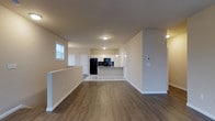 This two-story space features a clean, modern design with an open-concept layout that connects the main living area to the kitchen seamlessly. The main level is finished with rich, dark hardwood-style flooring that contrasts nicely against the neutral beige and white walls, creating a warm yet contemporary atmosphere. Recessed and flush-mount ceiling lighting illuminates the space evenly throughout.

The kitchen stands out as a central feature, offering white cabinetry that extends to the upper walls, a kitchen island or peninsula with a countertop overhang ideal for seating, and a full suite of black appliances including a refrigerator, microwave, and range. The kitchen transitions to lighter flooring, distinguishing it from the main living area.

A staircase with a half-wall railing is visible, indicating access to the second floor. Natural light enters through a window near the stairwell, brightening the space. The open floor plan, neutral color palette, and modern finishes throughout give the space a versatile and move-in-ready feel, well-suited for a variety of furnishing styles and uses.