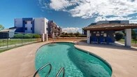 Located at 1469 Zepol Rd in Santa Fe, New Mexico, this outdoor amenity area serves as a communal recreational space set against the backdrop of the surrounding multi-story residential buildings. The centerpiece of the space is a generously sized, kidney-shaped swimming pool with clear turquoise water, accessible via stainless steel handrails and marked with depth indicators for safety. The pool is complemented by a broad, curved concrete deck that wraps around its perimeter, providing ample room for lounging and movement. A covered cabana structure anchors one side of the pool area, featuring a tile roof supported by painted concrete columns, offering a shaded retreat with seating for residents. Multiple blue lounge chairs are positioned throughout the deck and surrounding grassy areas, inviting relaxation in the abundant New Mexico sunshine. The grounds are enclosed by a wrought iron fence, ensuring privacy and security, while patches of maintained lawn add greenery to the hardscape. The open layout of the space affords sweeping views of the expansive New Mexico sky, with the surrounding architecture providing a sense of community. The overall ambiance is bright, open, and resort-inspired, ideal for outdoor recreation and leisure.