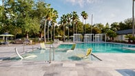 Located in the Citrus Village community of Tampa, Florida, this single-story outdoor amenity facility offers residents a well-appointed recreational destination. At its centerpiece is a generously sized swimming pool with crystal-clear turquoise water, surrounded by a spacious sun deck lined with lounge chairs — all framed by towering palm trees and mature tropical foliage that provide a serene, resort-like atmosphere.

Adjacent to the pool is a covered pavilion that serves as the social hub of the space. The pavilion features polished concrete flooring, a shiplap-style white ceiling with modern ceiling fans, and open-air walls supported by square columns that allow for seamless indoor-outdoor flow with direct sightlines to the pool. Seating includes multiple round and rectangular tables paired with colorful contemporary chairs in navy and yellow-green tones.

A built-in wet bar or kitchenette anchors one side of the pavilion, complete with pendant lighting, bar-height seating, and a dark faucet fixture — ideal for entertaining. A covered walkway connects additional areas of the facility, providing shaded passage with Adirondack-style seating tucked alongside the pool perimeter. The overall design balances functional amenities with a bright, casual Florida aesthetic.