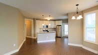 This single-floor space showcases an open-concept layout that seamlessly integrates the kitchen and living areas, creating a bright and functional environment. The kitchen serves as the focal point, featuring crisp white cabinetry that extends to the upper walls, providing ample storage. Stainless steel appliances, including a refrigerator, range, and microwave, lend a modern, polished aesthetic. A central island offers additional counter space and serves as a natural gathering point. Pendant lighting fixtures hang above the island, adding warm, focused illumination that complements the overall design. The neutral taupe-toned walls provide a warm, cohesive backdrop throughout the space, while rich, dark hardwood-style flooring runs continuously across the entire area, grounding the open layout with a sense of warmth and continuity. White baseboards offer a clean contrast against the flooring. Natural light filters in through a door with a window, brightening the space. A hallway visible in the background suggests additional rooms beyond, contributing to the overall functional flow of this well-appointed single-floor residence.